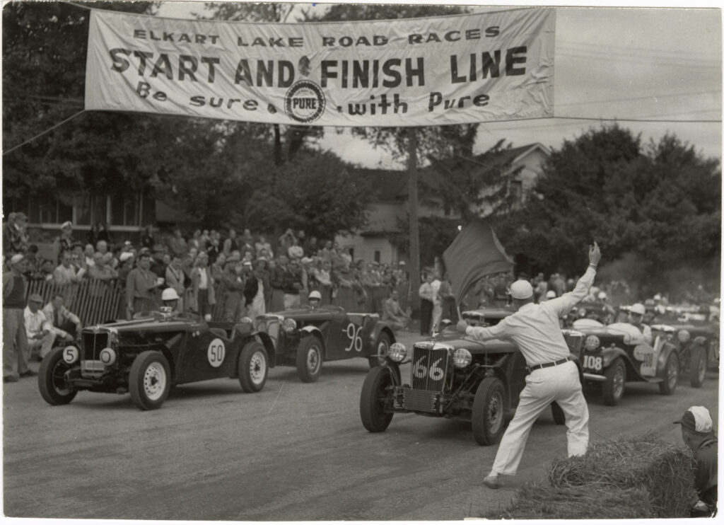 A black and white photo of a man waving a start flag to a car race. A banner above reads Elkart Lake Road Races. Start and Finish Line. Be sure. With Pure.